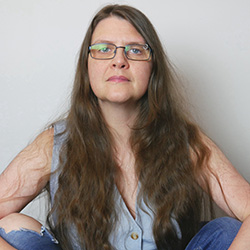 Photo alt-text: A self-portrait of San, a 52-year-old white person with long brown hair, blue eyes, an eyebrow piercing and blue-rimmed metal glasses. They're wearing a low-cut sleeveless waistcoat and jeans with a hole in one knee, and sit cross-legged with arms around their knees. San looks directly into the camera with an open expression.
